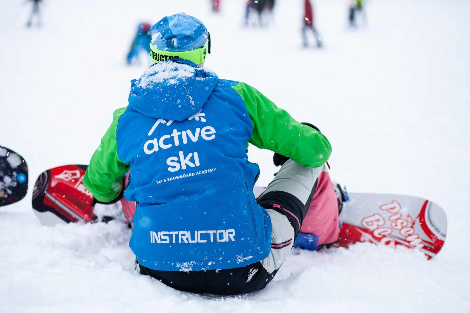 Zuzy sedí chrbtom k fotoaparátu na snehu v bunde inštruktorky, vedľa nej snowboard s nápisom Píšem Blog